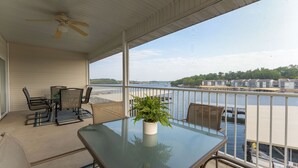 A balcony with a glass table and chairs, a potted plant, and a view of a waterfront with buildings and a dock. - In the Middle of it All -2 Pools,Grill,Fishing,Boat Launch,Boat rental next door (Osage Beach)