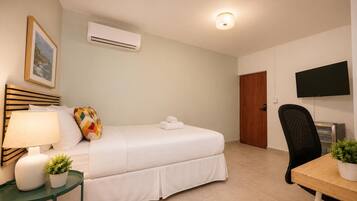 A hotel room with a bed, bedside table with a lamp, a TV, a desk with a chair, and an air conditioning unit.