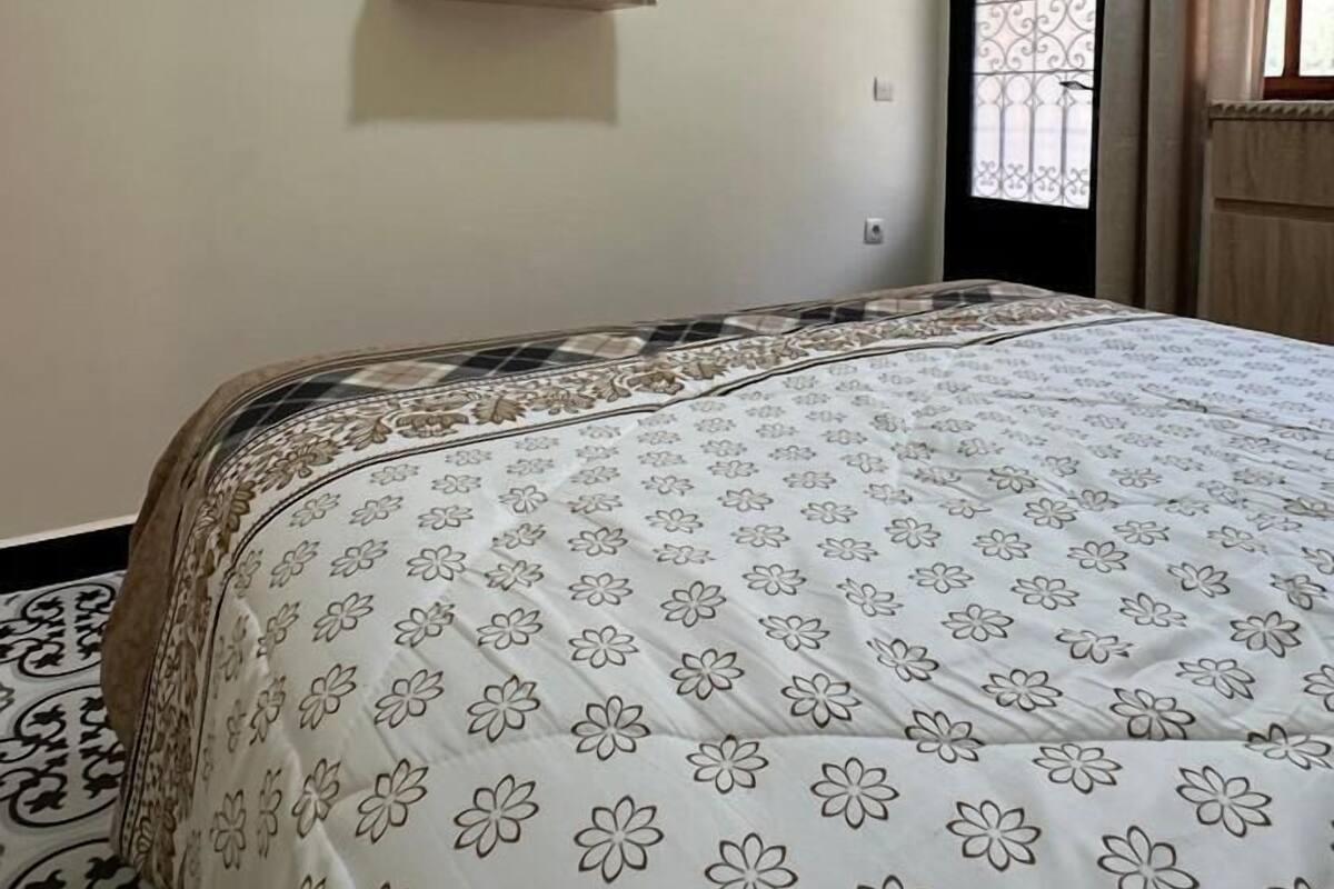 A bedroom with a bed, a mounted TV, a small shelf, and a window with curtains.