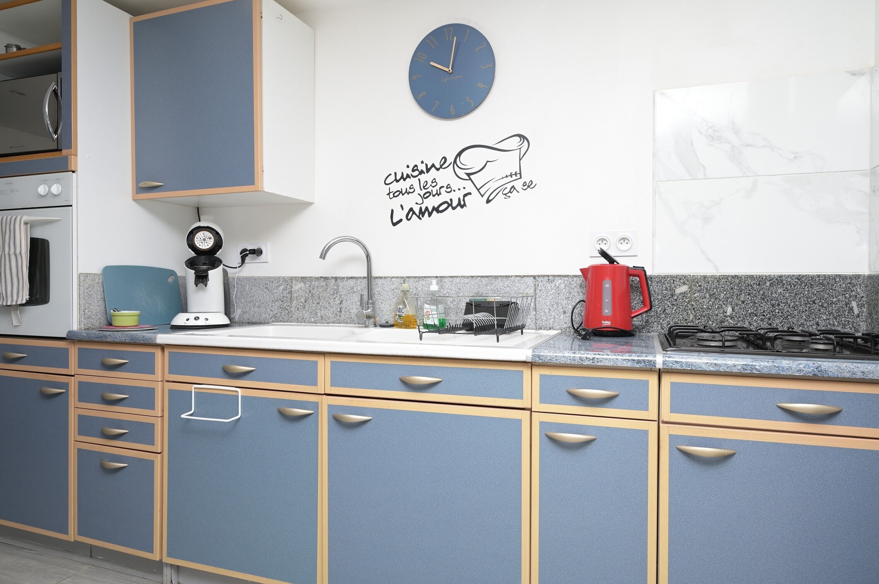 Een keuken met blauwe kastjes, een wit aanrecht, een klok, een koffiezetapparaat en een fornuis.