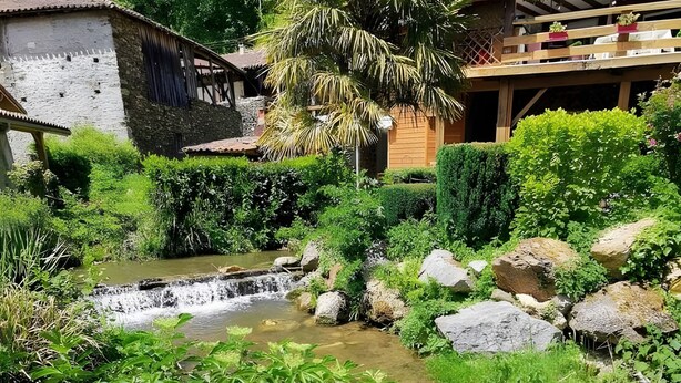 Een waterval in een weelderige tuin, met een houten huis en een stenen gebouw op de achtergrond.