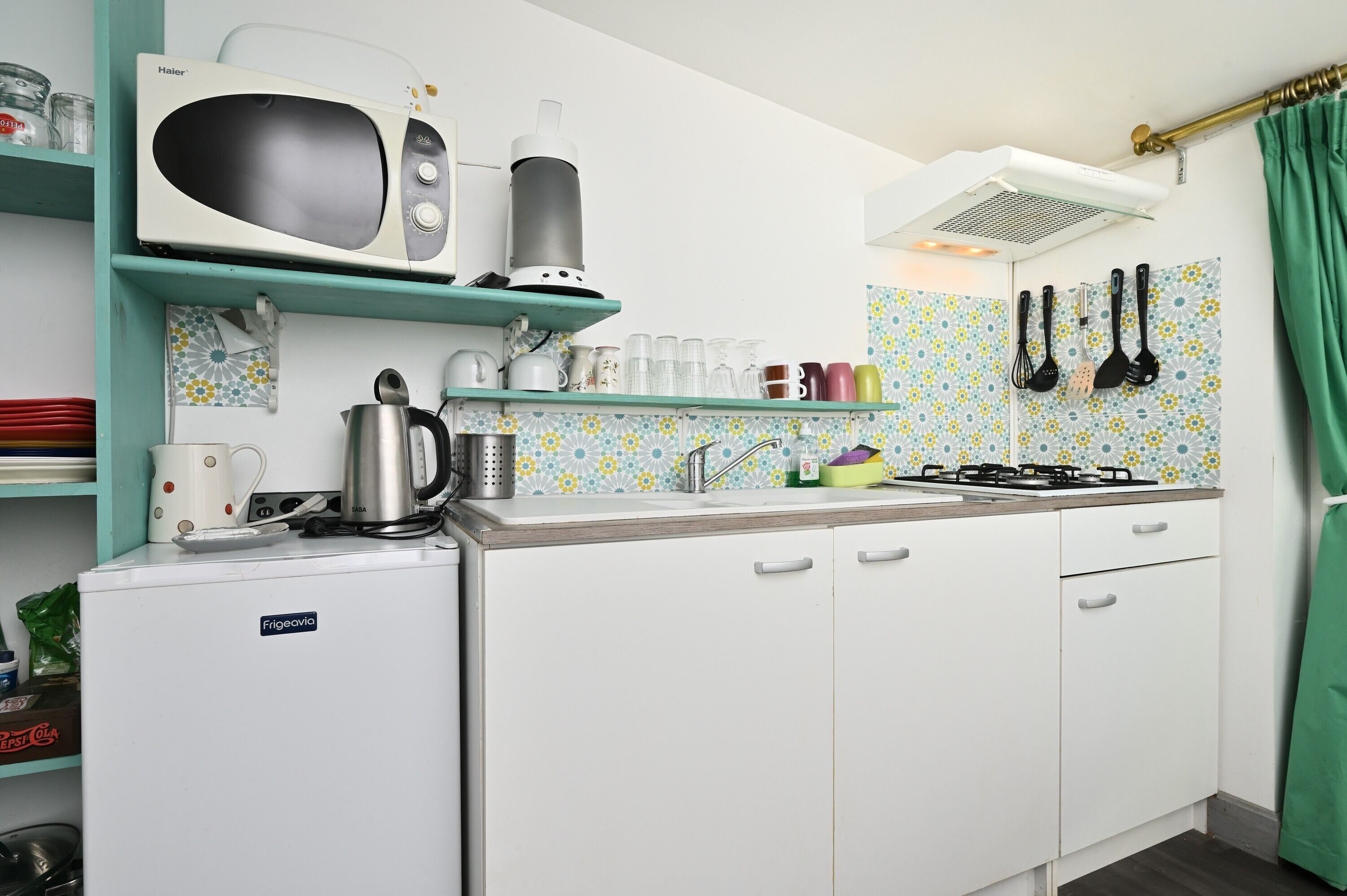 A compact kitchen with a microwave, kettle, and stove. There are shelves with various kitchen items and a patterned backsplash.