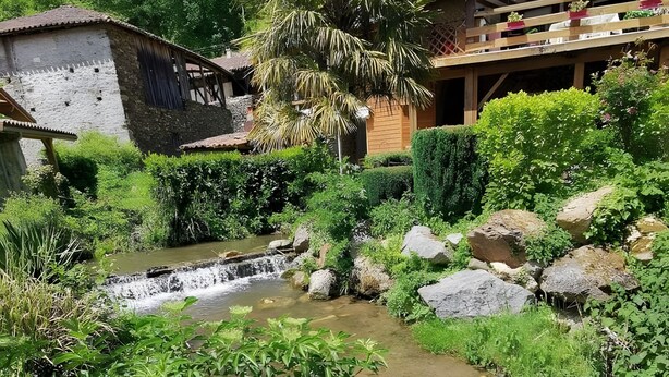 Un edificio rústico con un balcón de madera, una cascada pequeña y abundante vegetación.