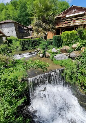 Un edificio rústico con un balcón de madera, una cascada pequeña y abundante vegetación.