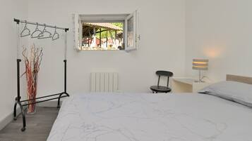 A bedroom with a bed, a radiator, a window with a view, a hanging clothes rack, and a bedside table with a lamp.