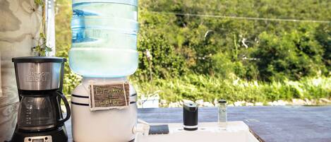 Un distributeur d’eau, une machine à café et un petit récipient sont posés sur une table, offrant une vue sur un paysage verdoyant.