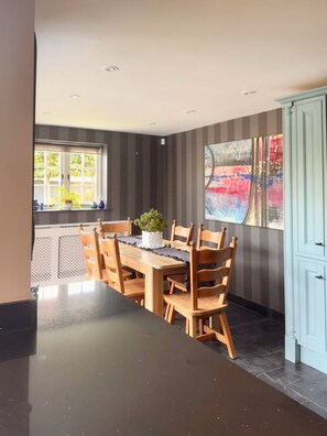 Un'area pranzo con un tavolo e sedie in legno, una finestra con piante in vaso e un muro decorato con un dipinto astratto dai colori vivaci.