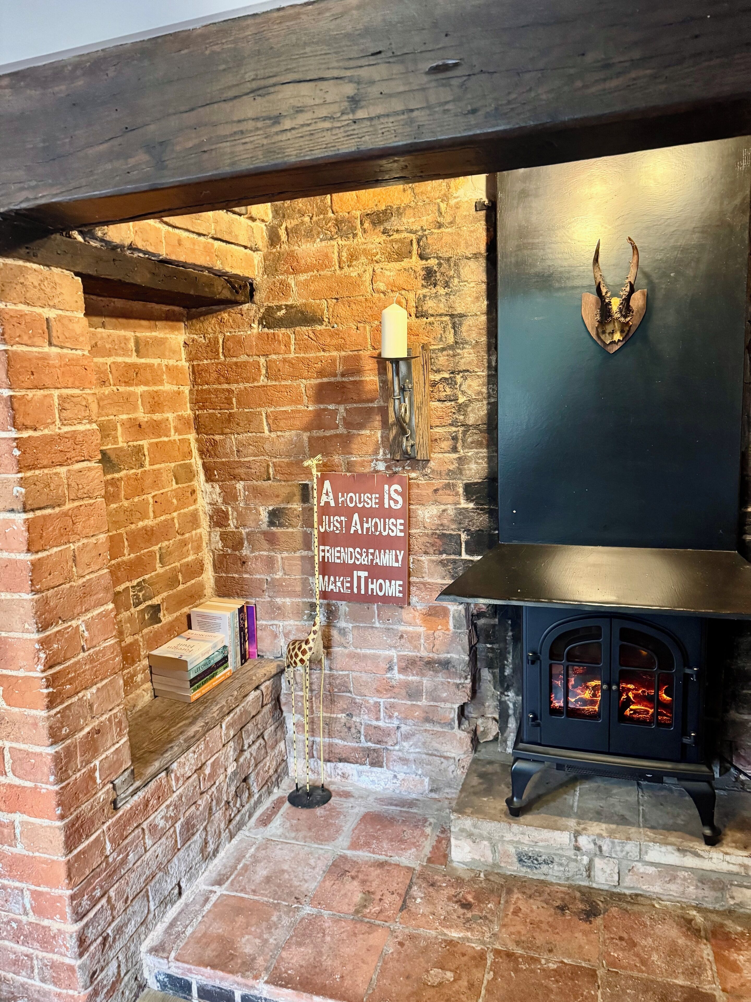 A cozy fireplace area with a brick wall, a wooden mantel, a sign stating 'A HOUSE IS JUST A HOUSE FRIENDS&FAMILY MAKE IT HOME', and a decorative deer head.