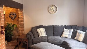 Una sala de estar acogedora con un sofá seccional, un pequeño reposapiés, un reloj de pared decorativo, una lámpara colgante y una pared de ladrillo con un adorno con forma de corazón.