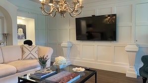 Una sala de estar con un sofá color beige, una mesa de centro de cristal con libros, una araña y un televisor de pantalla plana montado en la pared.