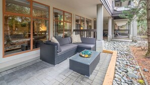 Un espace extérieur aménagé, abrité, avec un canapé gris et une table basse en osier, de grandes fenêtres et un chemin en pierre.