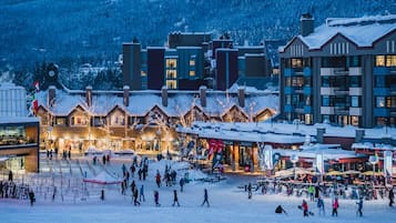 Una piazza innevata con edifici, tra cui un lodge con una torre dell'orologio, e persone che camminano e sciano.
