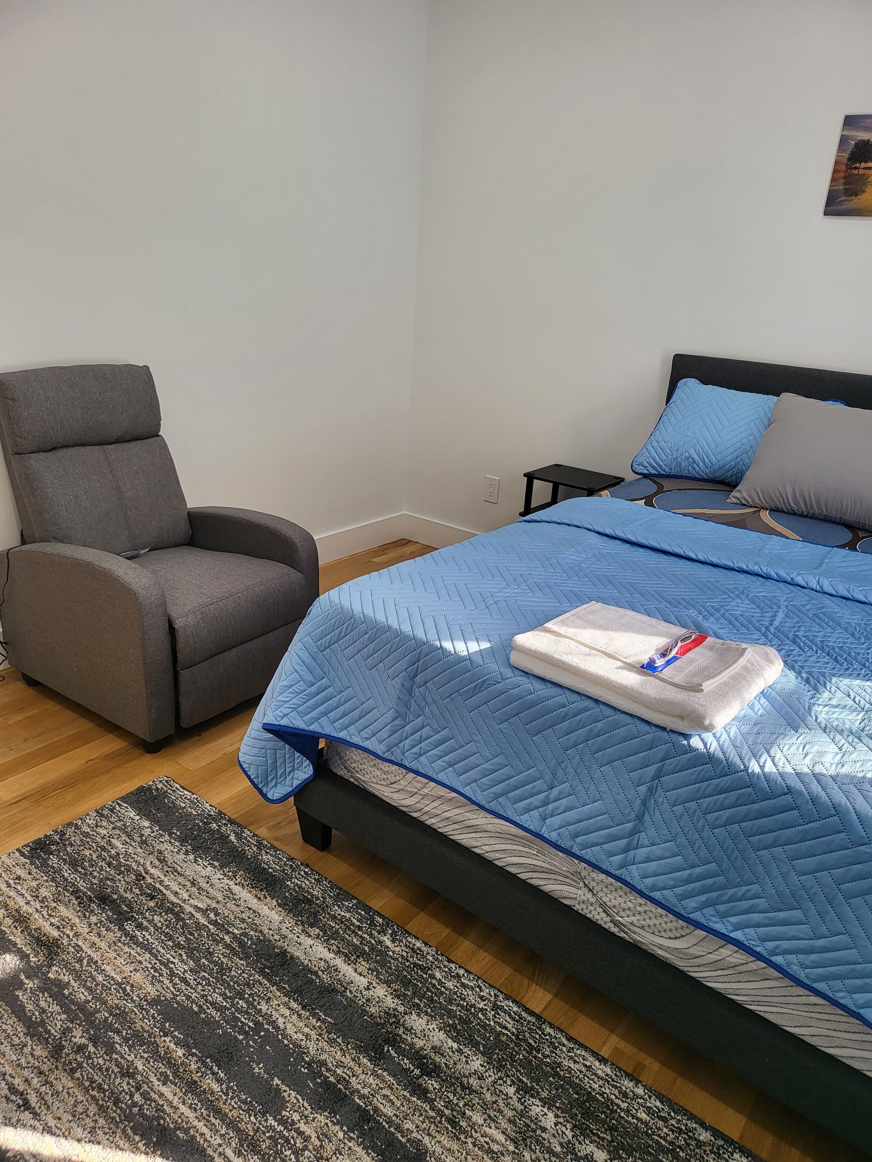 Una camera da letto con un letto, una poltrona grigia, un comodino e un quadro appeso al muro.