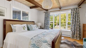A bedroom with a large bed, wooden beams, a window with curtains, and a view of greenery outside.