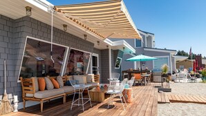 A covered outdoor patio with a wooden deck, a seating area with a sofa and chairs, a dining table, and a barbecue grill.