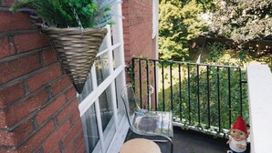 A small patio area with a brick wall, a hanging plant, a clear plastic chair, a small round table, and a metal fence.