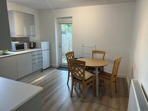 Una cocina compacta con microondas, hervidor y armarios. Hay una mesa de comedor con sillas y vistas al exterior a través de una puerta de cristal.