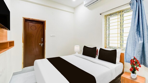 Ein Hotelzimmer mit Bett, Nachttisch, Lampe, einem Fenster mit Vorhängen, einer Klimaanlage und einer Tür.