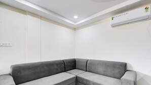 Ein Eckzimmer mit grauem Ecksofa, weißen Wänden, einer eingelassenen Deckenleuchte und einer Wand-Klimaanlage.