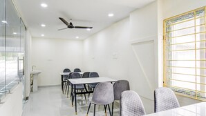Ein Konferenzraum mit weißem Tisch, grauen Stühlen, einem Deckenventilator und einem Fenster mit Gittermuster.