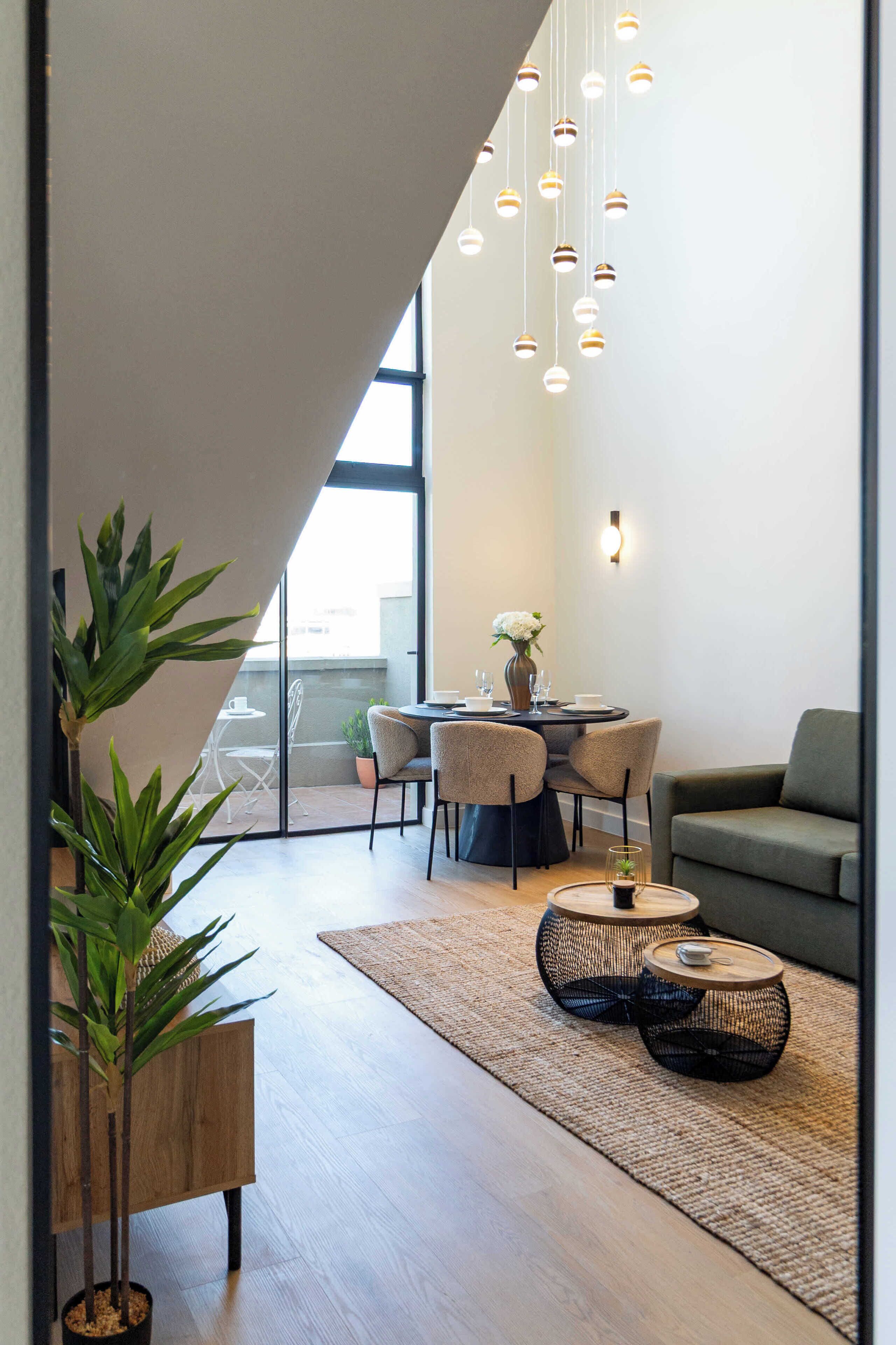 A modern living room with a dining area, featuring a large window, hanging lights, and a green sofa.