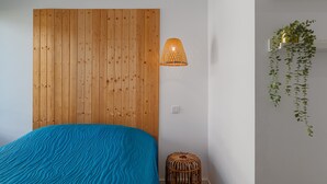 A bedroom with a wooden headboard, a blue textured bedspread, a wicker stool, a hanging plant, and a woven pendant light.