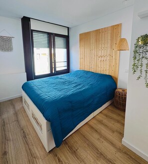 A bedroom with a bed, wooden floor, a window with blinds, a wall-mounted lamp, and a hanging plant. - Casa do Sal – Your Refuge Near the Sea (Troia)