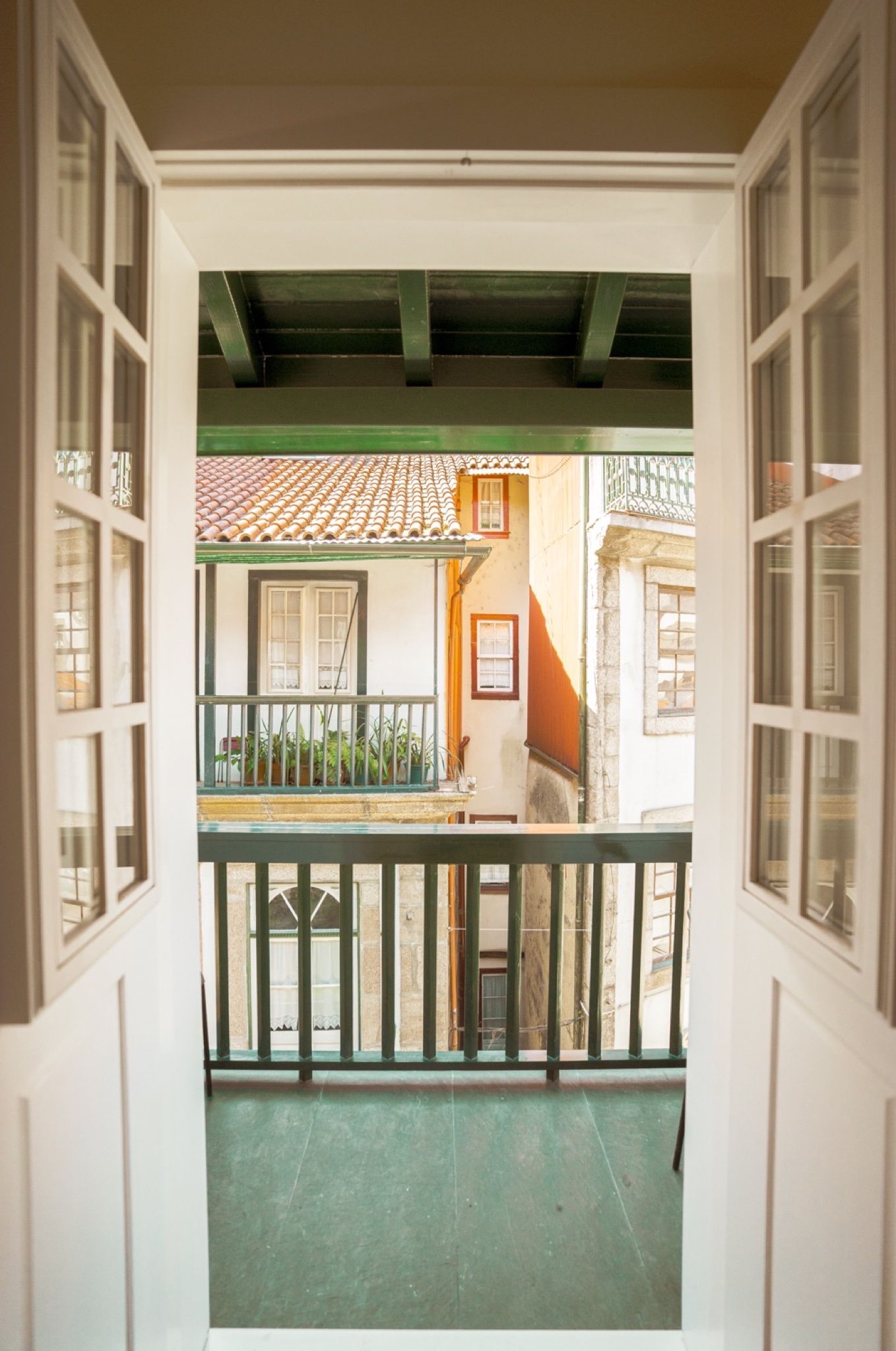 Une vue à travers une porte ouverte donnant sur un balcon doté d’un toit en tuiles, d’une petite table et de plantes en pot.