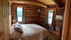 Ein gemütliches Blockhaus-Zimmer mit einem Bett, zwei mit Vorhängen versehenen Fenstern, einer Lampe und einer Klimaanlage.