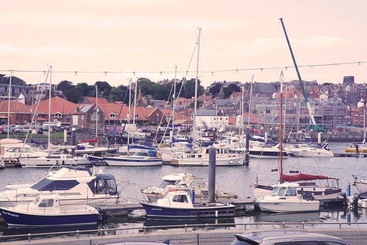 A marina with multiple boats docked, surrounded by buildings and a hilly landscape in the background.