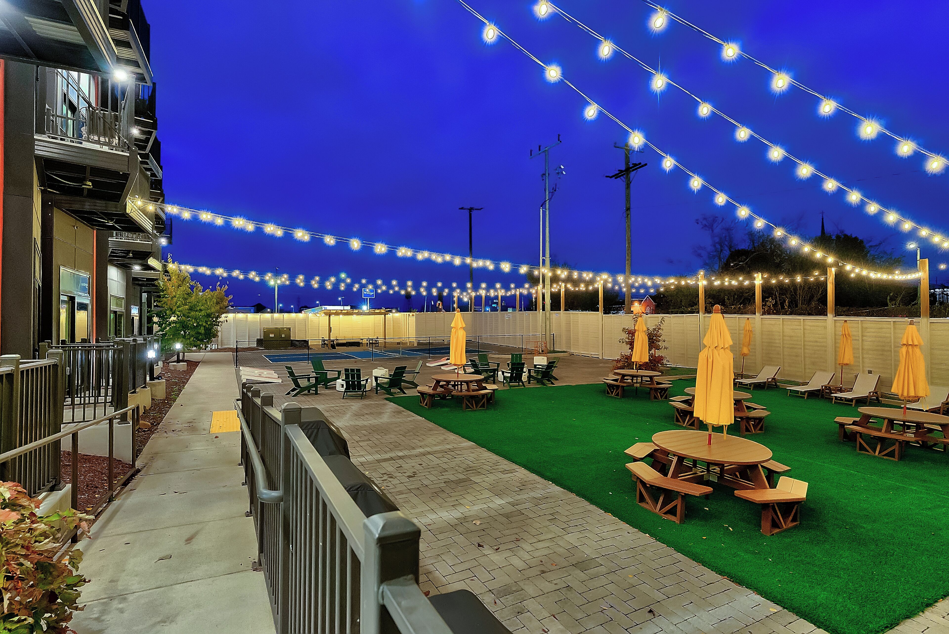 Un patio al aire libre con luces colgantes, césped artificial verde y mesas y sillas de madera.