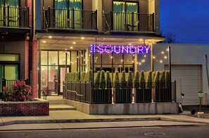 Ingresso di un hotel con un neon che riporta la scritta "SOUNDRY", un cancello e un piccolo giardino con piante in vaso.