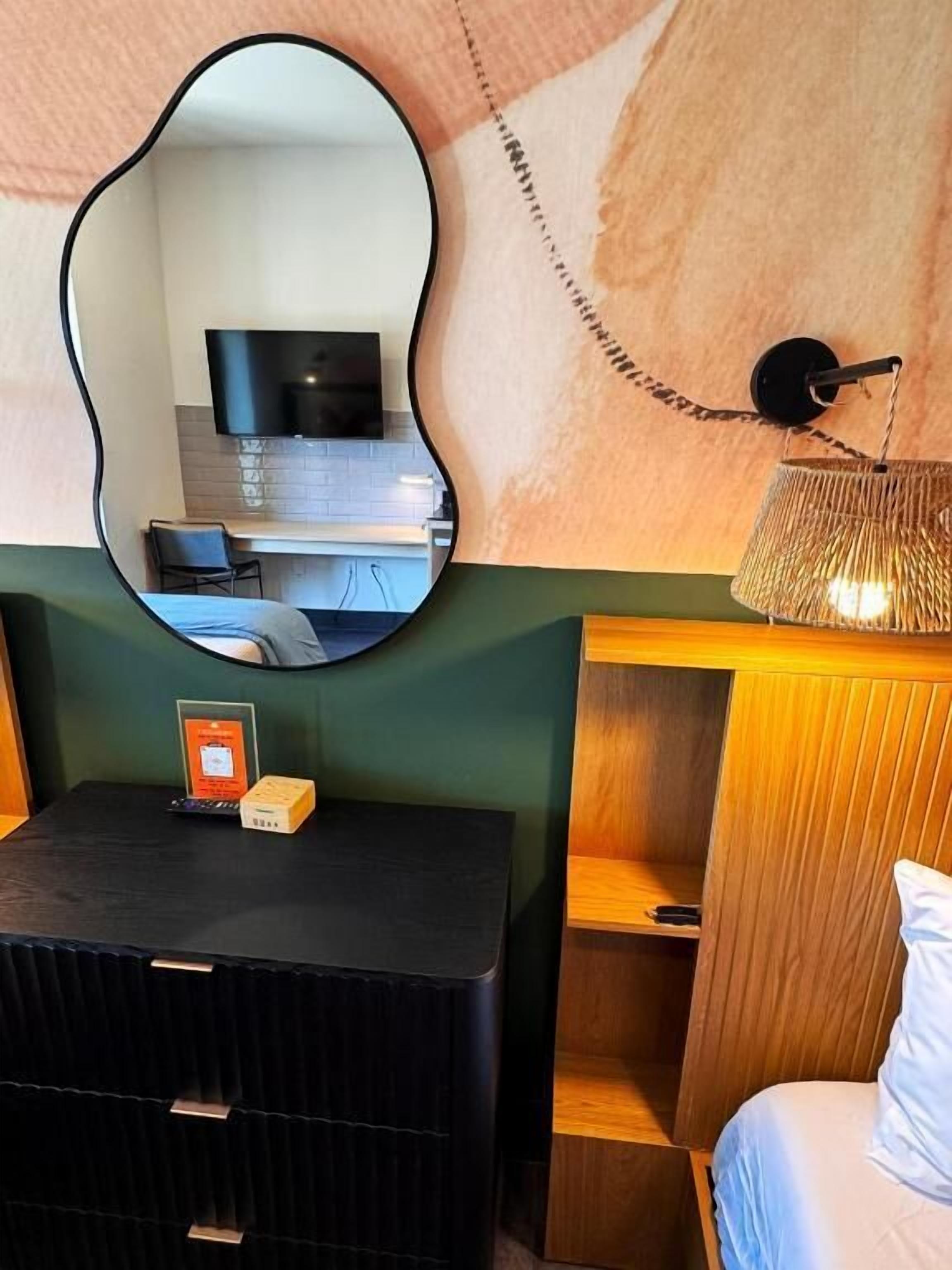 Habitación de hotel compacta con pared espejada, un pequeño escritorio con silla, una cama y un televisor montado en la pared.