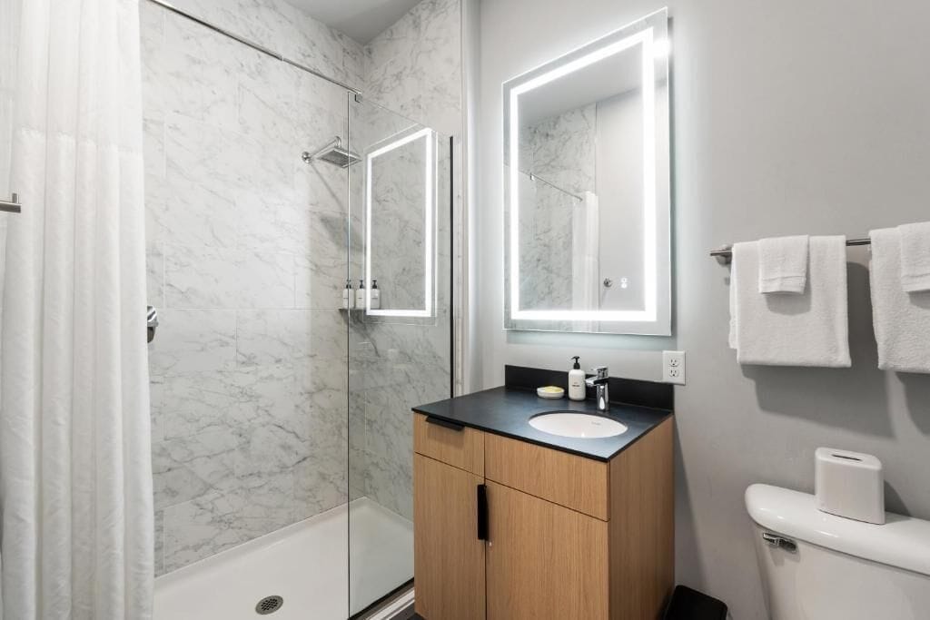 A modern bathroom with a glass shower, marble walls, a wooden vanity, and two illuminated mirrors.
