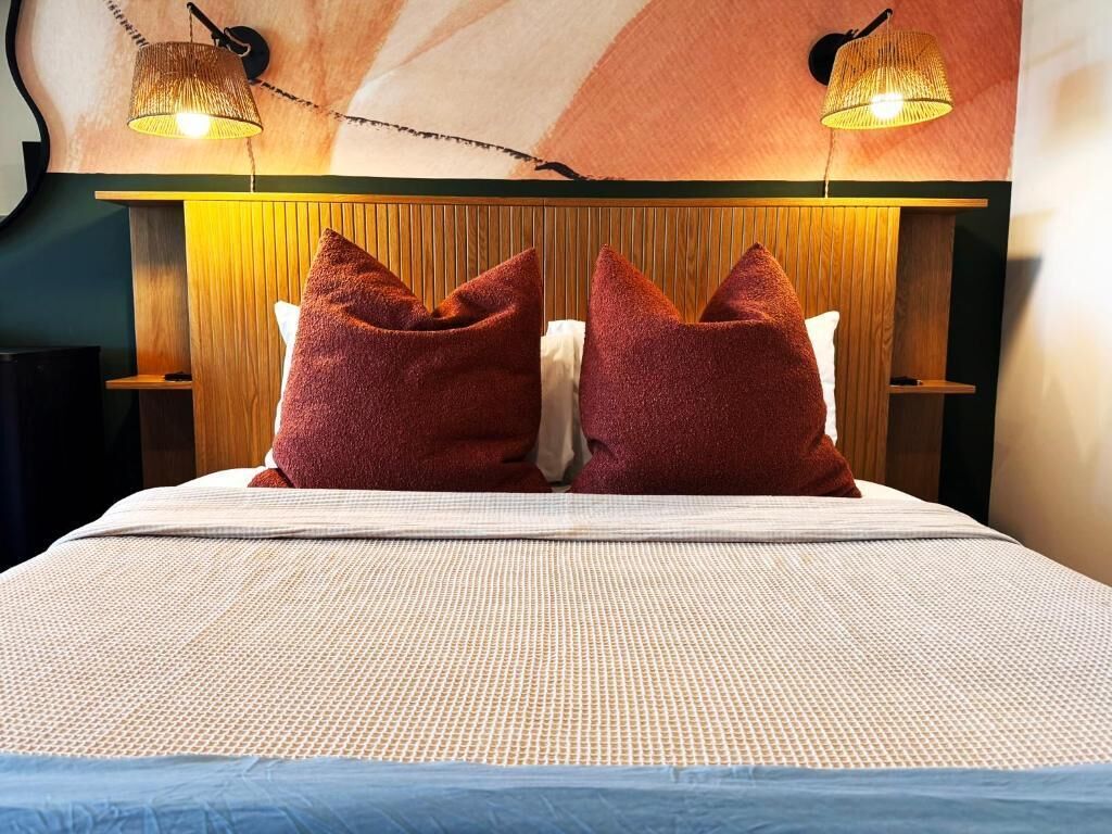 Cama com colcha branca texturizada, duas almofadas vermelhas e cabeceira de madeira com luminária trançada acima.
