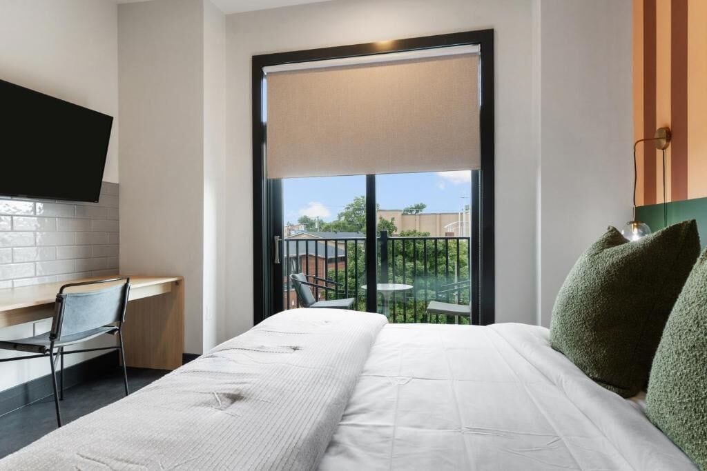 Habitación de hotel con cama, escritorio, silla, televisor montado en la pared y balcón con mesa y sillas.