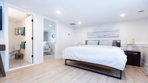 A modern bedroom with a large bed, a bedside table with a lamp, and a view of the bathroom through an open door. - Modern Renovated Retreat w/ Pool in North Cape May (Lower Township)