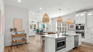 A spacious kitchen with white cabinetry, a large island with a sink, and a dining area with wooden chairs. - Sleeps 34! 2 Homes, Private Pools, Firepit, Golf Cart, Walk to Beach Seagrove San Juan by Avantstay (Santa Rosa Beach)