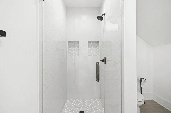 Une douche fermée par des parois de verre, avec un sol carrelé de carreaux blancs et une porte dotée d’une poignée.