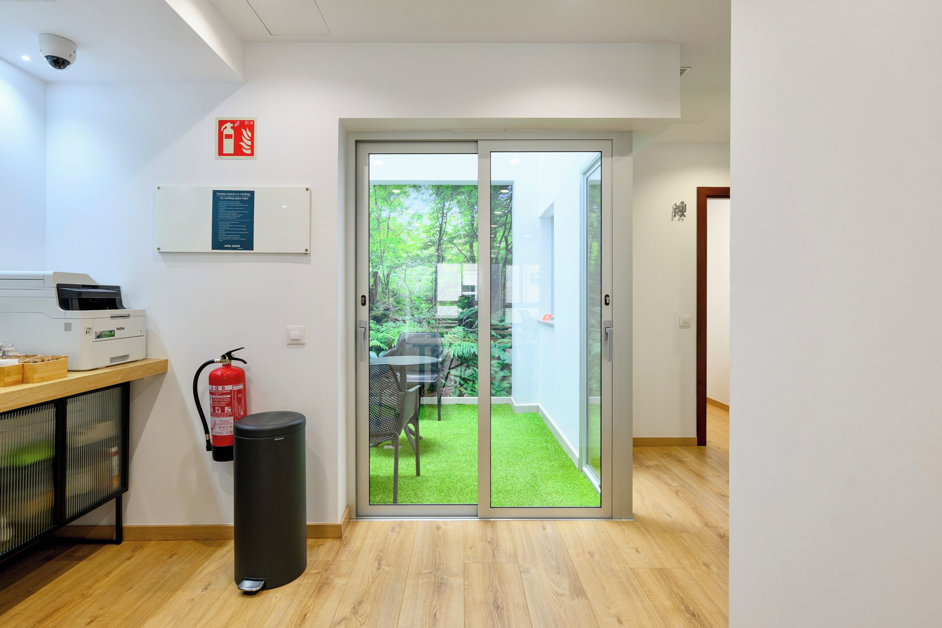 Habitación de hotel con puerta de vidrio corredera que da a un jardín, extintor, cesto de basura y escritorio con impresora.
