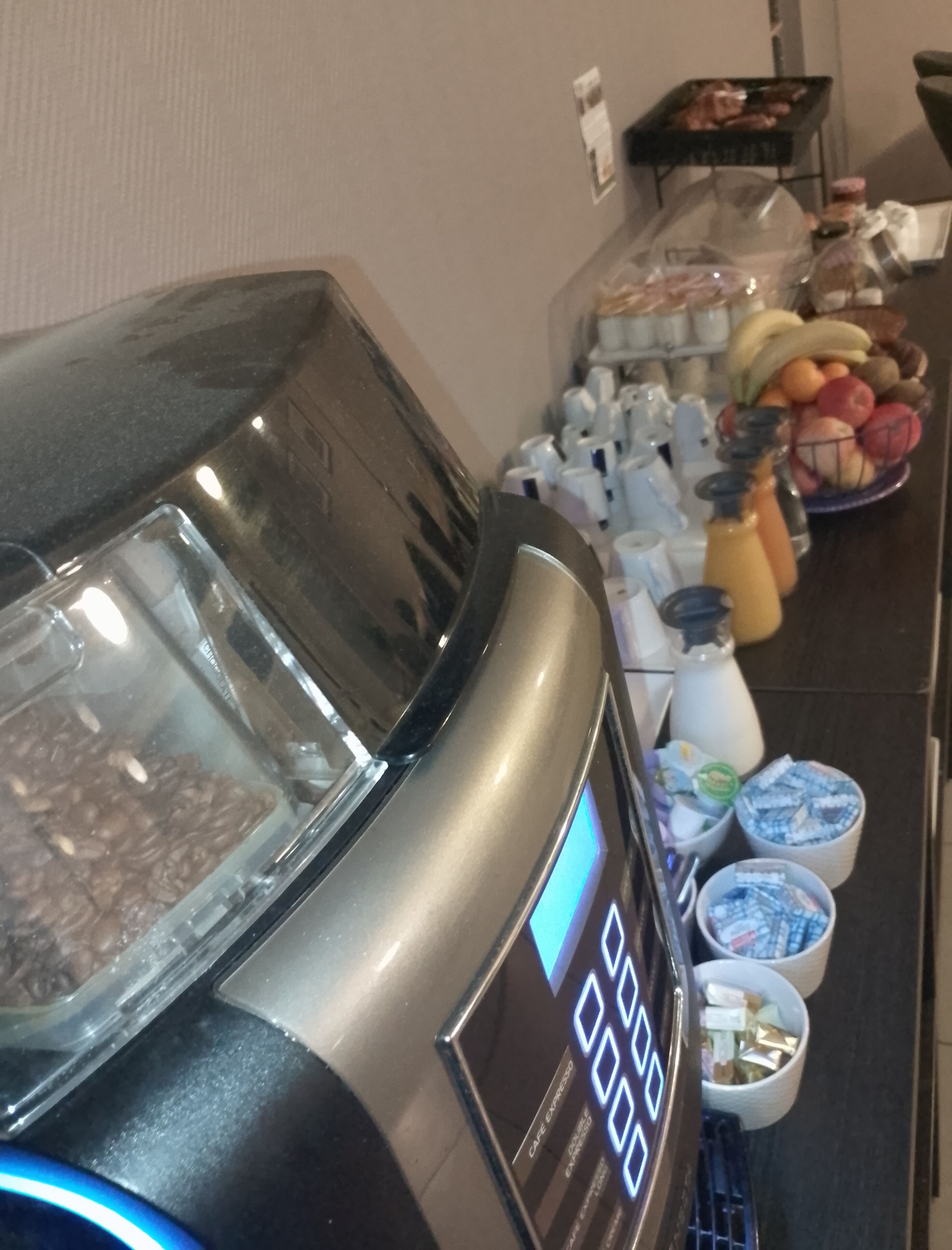 A coffee machine with a digital display, a glass container with nuts, and a fruit bowl on a counter.