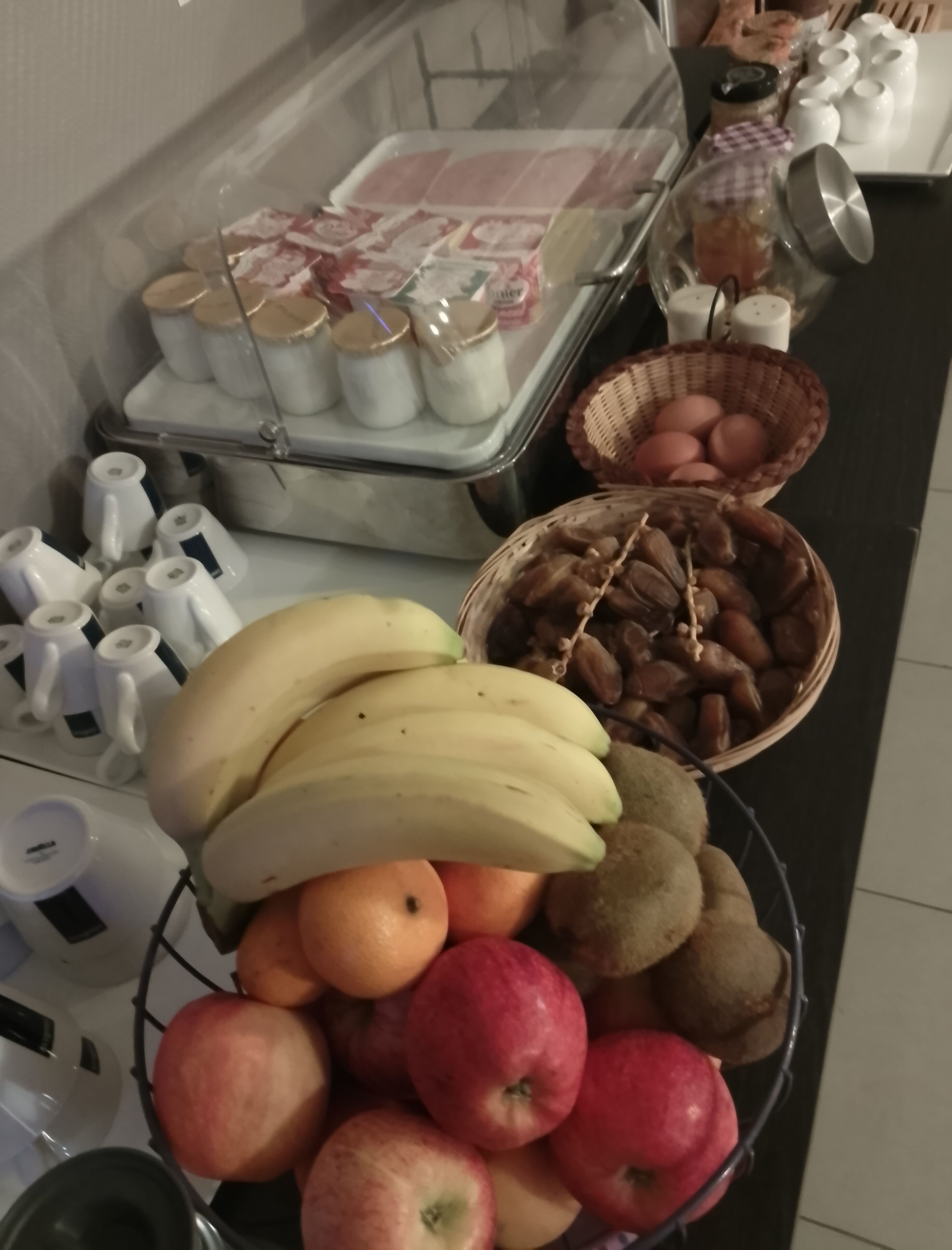 A kitchen counter with a fruit bowl containing bananas, apples, and kiwis, a basket of nuts, and a container with jars of condiments.