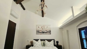 A bedroom with a wooden ceiling beam, a bed with a quilted bedspread, a nightstand with a lamp, and a window with curtains. - Exclusive Family Vacation: High-Quality, Stylish Apartment in Milan, 10 Minutes from Gardaland (Venetien)