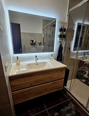 Un baño moderno con un espejo grande, un lavamanos blanco, cajones de tocador de madera y una mampara de ducha de vidrio.