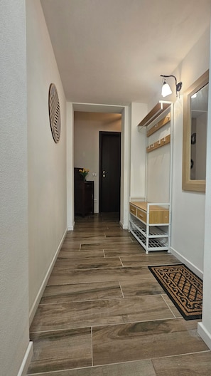 Un pasillo con piso de madera, una pared blanca con un panel decorativo, una puerta y un estante blanco con un espejo.