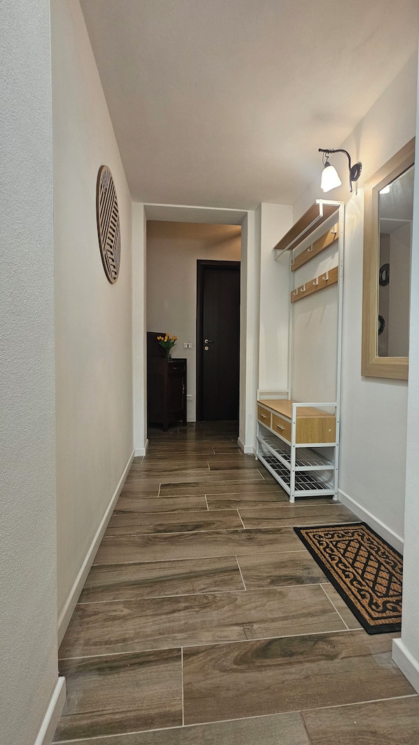 A hallway with a wooden floor, a white wall with a decorative panel, a door, and a white shelf unit with a mirror.