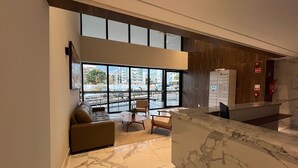 A modern lobby with a marble reception desk, a seating area with a sofa and chairs, large windows offering a view of the street, and a Vertech information kiosk. - Apartamento Noroeste (Brasília)