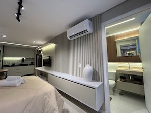 A modern hotel room with a bed, a TV, a minibar, a bathroom with a sink and mirror, and a kitchen area with a refrigerator and stove. - Apartamento Noroeste (Brasília)