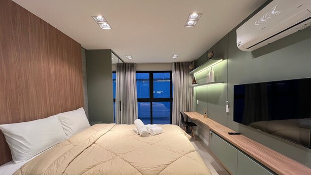 A modern hotel room with a large bed, a desk, a flat-screen TV, and a view of the cityscape through the window. - Apartamento Noroeste (Brasília)