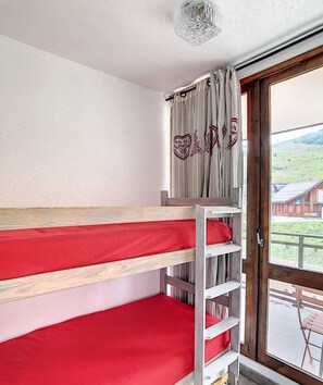 Ein Schlafraum mit Etagenbett, einer Holzlade, Vorhängen an einem Fenster und Blick auf ein Gebäude.